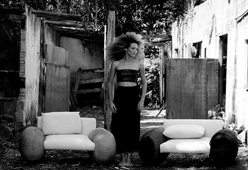 Mujer posando entre dos sillones tapizados con brazos tejidos, en un entorno rústico al aire libre. Diseño de mobiliario artesanal en contraste con arquitectura deteriorada.